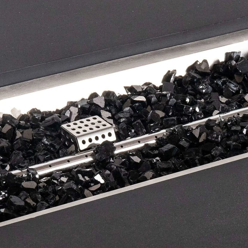 Detailed close-up of glossy black fire glass inside the black Flame Havens Refuge Series 56 x 30 x 24-inch rectangular GFRC concrete propane fire pit, showing the stainless steel linear burner and perforated ignition cover within the recessed burner pan.
