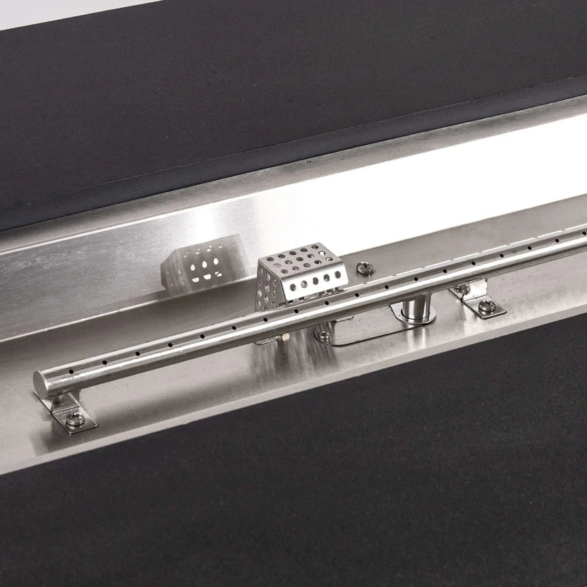 Tight close-up of the Flame Havens Refuge Series propane fire pit’s stainless steel linear burner and CSA ignition system, including evenly spaced burner ports, mounting brackets, wiring, and perforated protective cage set inside the black GFRC concrete surround.