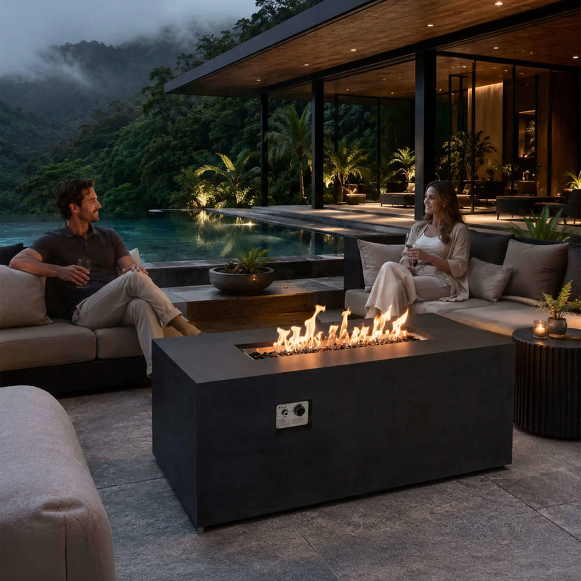 Flame Havens Refuge Series 56 x 30 x 24-inch black rectangular GFRC propane fire pit lit over tempered black fire glass on a luxury poolside patio. A couple relaxes with drinks beside the fire, surrounded by modern outdoor seating, tropical greenery, and misty mountain views.