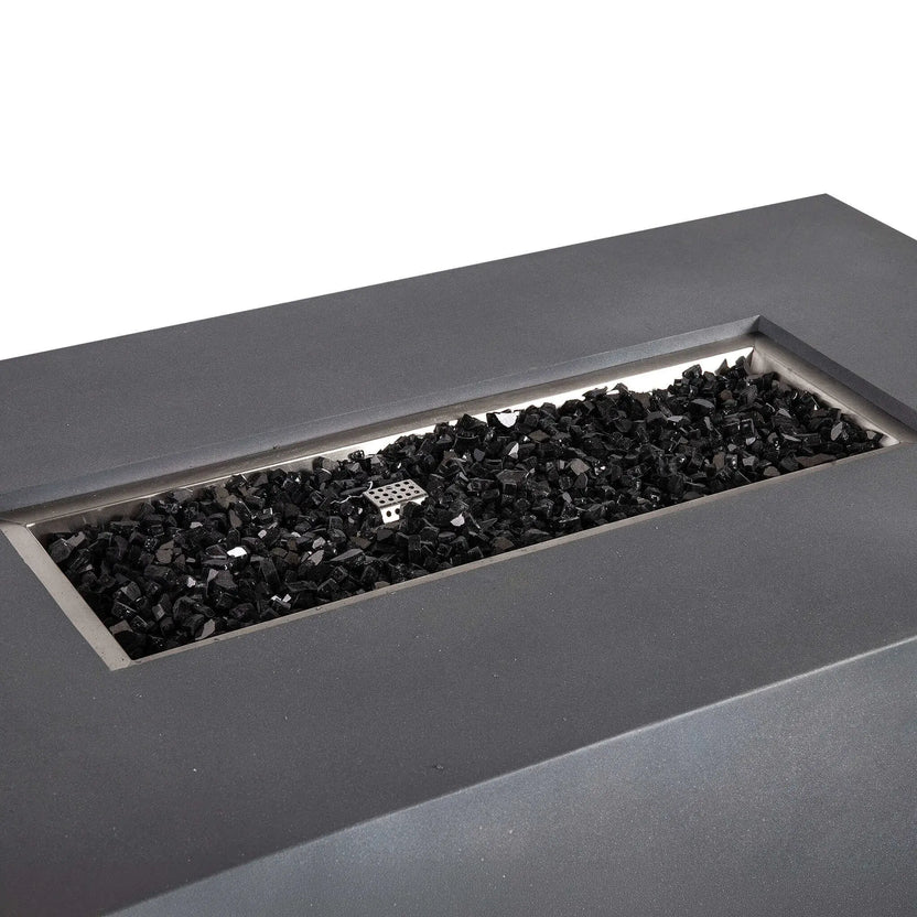 Angled close-up of the Flame Havens Refuge Series 56 x 30 x 24-inch rectangular propane gas fire pit in gray GFRC concrete. The recessed stainless steel burner pan is filled with reflective black fire glass, with the perforated ignition cover visible near the center against a clean white background.