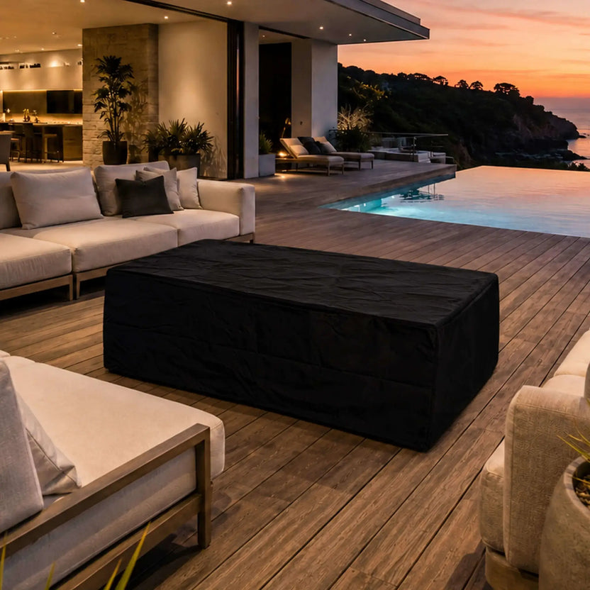Flame Havens Refuge Series 56 x 30 x 24-inch gray rectangular propane fire pit protected by a fitted black canvas fabric cover. The covered fire pit sits among cushioned outdoor seating on a modern poolside deck overlooking the coast at sunset, showing how it looks in a luxury patio setting when not in use.