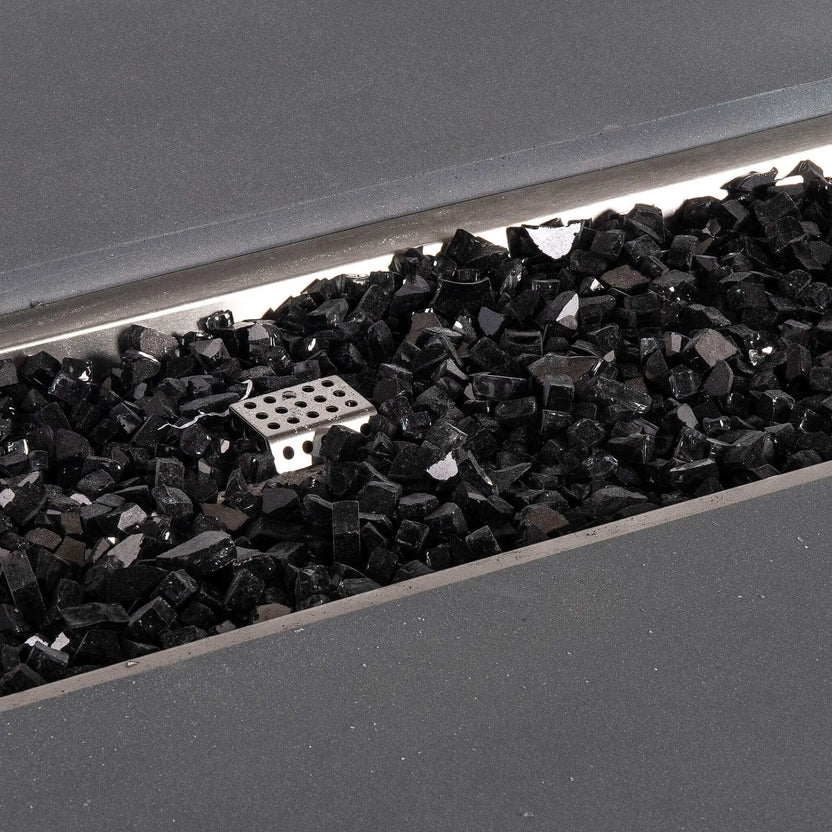 Detailed close-up of reflective black fire glass inside the Flame Havens Refuge Series 56 x 30 x 24-inch gray GFRC concrete propane fire pit. The polished glass pieces fill the rectangular stainless steel burner pan, surrounding the small perforated ignition housing beneath the wide concrete ledge.