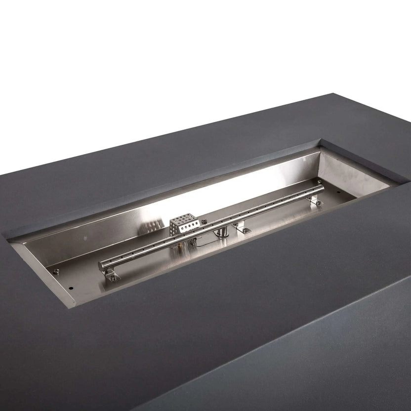 Angled close-up of the stainless steel burner assembly inside the Flame Havens Refuge Series 56 x 30 x 24-inch gray rectangular propane fire pit. The empty burner pan reveals the linear burner tube, evenly spaced flame ports, mounting brackets and perforated CSA ignition component.