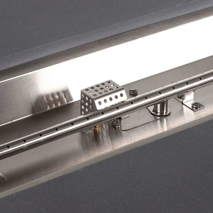Detailed view of the CSA ignition and stainless steel burner inside the Flame Havens Refuge Series 56 x 30 x 24-inch gray GFRC propane fire pit. The close-up highlights the linear burner tube with individual flame ports, perforated ignition housing, mounting hardware and reflective stainless steel pan.