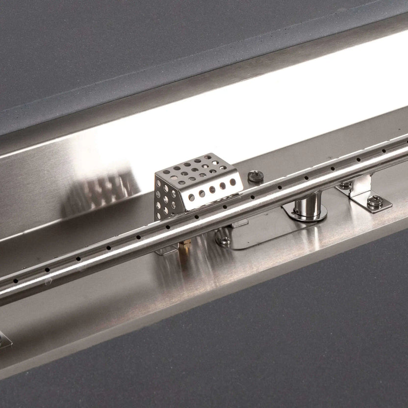 Detailed view of the CSA ignition and stainless steel burner inside the Flame Havens Refuge Series 56 x 30 x 24-inch gray GFRC propane fire pit. The close-up highlights the linear burner tube with individual flame ports, perforated ignition housing, mounting hardware and reflective stainless steel pan.