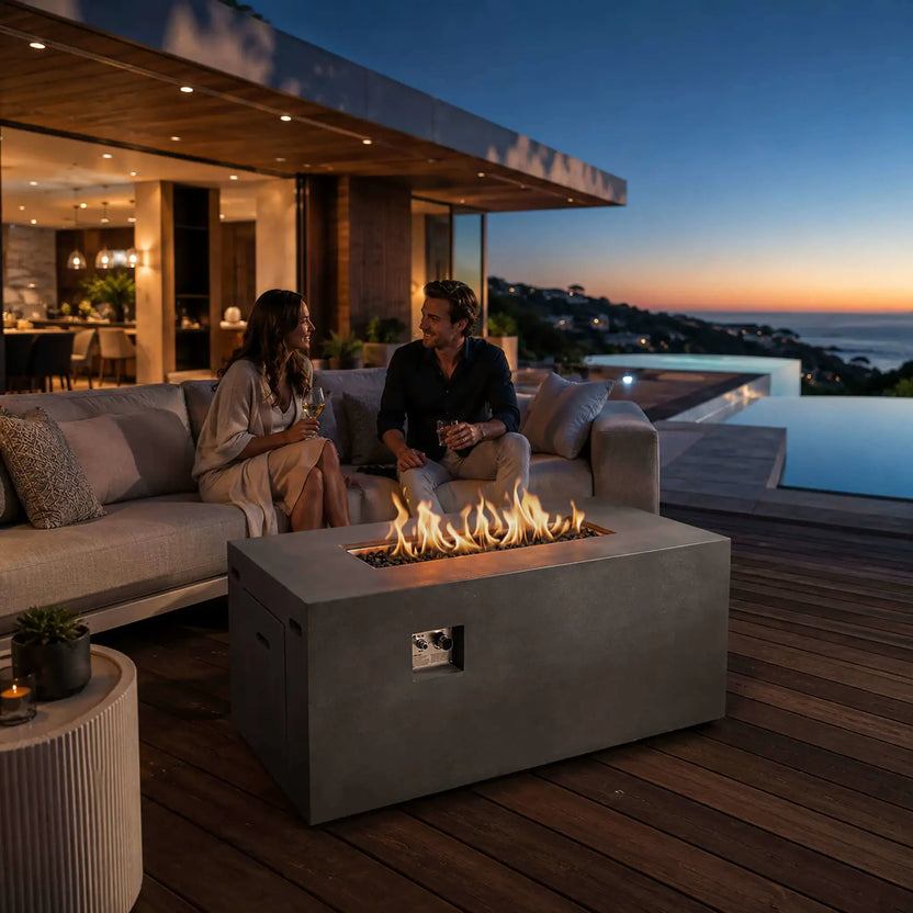 Flame Havens Refuge Series 56 x 30 x 24-inch gray rectangular propane fire pit glowing on a luxury coastal patio at sunset. A couple enjoys drinks on a cushioned sofa behind the fire pit as flames rise from tempered black fire glass. The front controls and hidden propane tank drawer are visible beside the modern pool and ocean view.