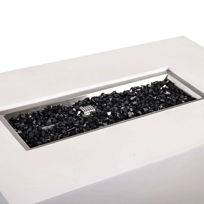 Angled close-up of the Flame Havens Refuge Series 56 x 30 x 24-inch rectangular propane gas fire pit in white GFRC concrete, showing the recessed stainless steel burner pan filled with black reflective fire glass and the central ignition cage.