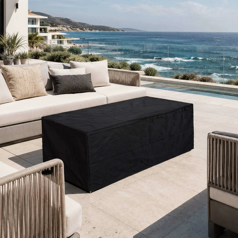 Flame Havens Refuge Series 56 x 30 x 24-inch rectangular white propane gas fire pit protected by a fitted black canvas fabric cover on a luxury coastal patio, surrounded by cushioned outdoor seating with a pool and ocean view in the background.