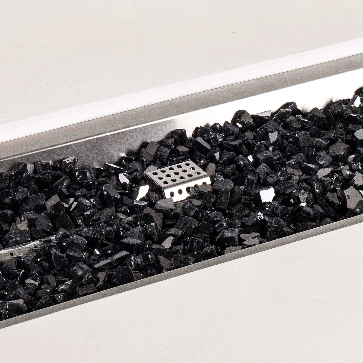 Detailed close-up of black reflective fire glass inside the stainless steel burner pan of a white Flame Havens Refuge Series 56 x 30 x 24-inch rectangular propane gas GFRC concrete fire pit, with the perforated ignition cage visible near the center.