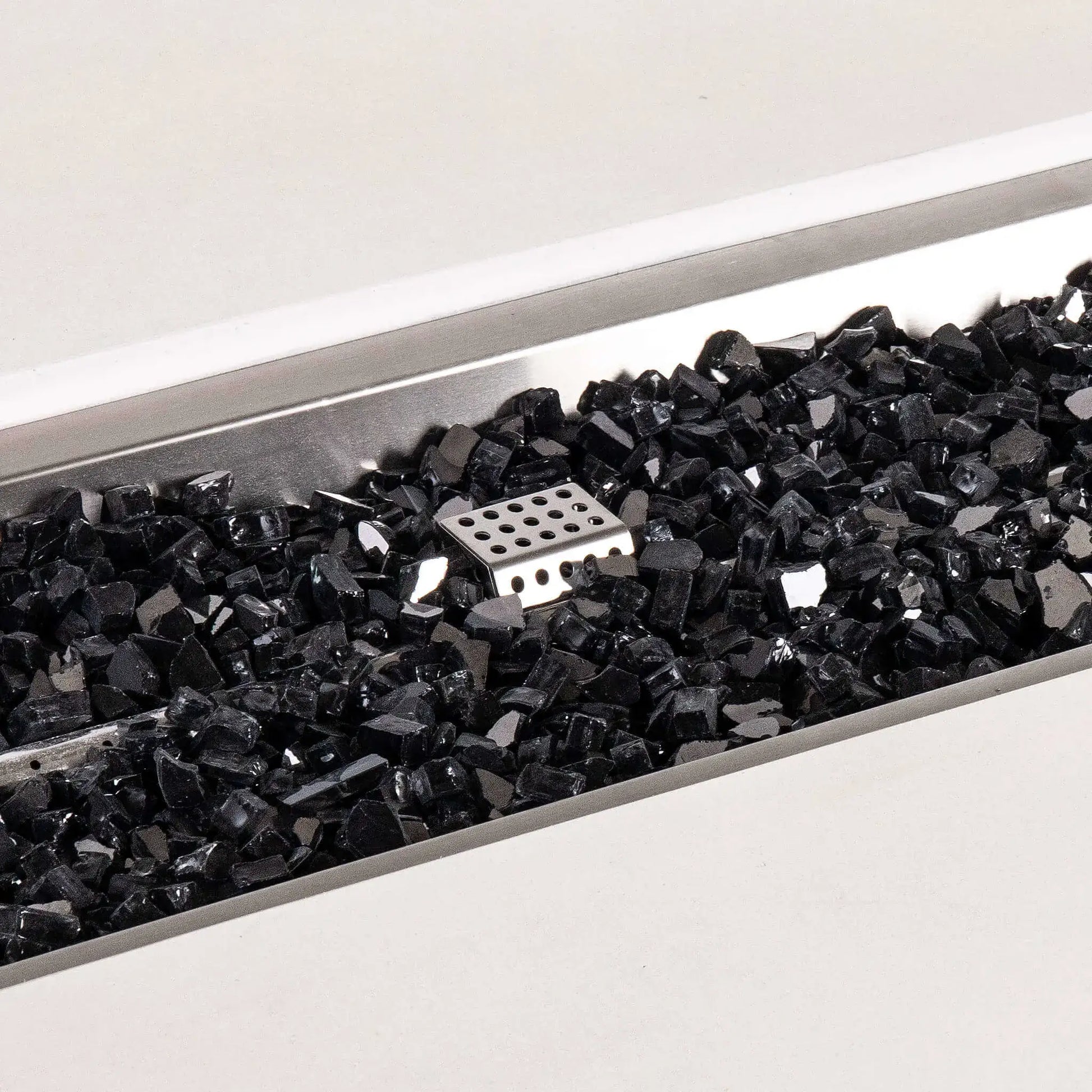 Detailed close-up of black reflective fire glass inside the stainless steel burner pan of a white Flame Havens Refuge Series 56 x 30 x 24-inch rectangular propane gas GFRC concrete fire pit, with the perforated ignition cage visible near the center.