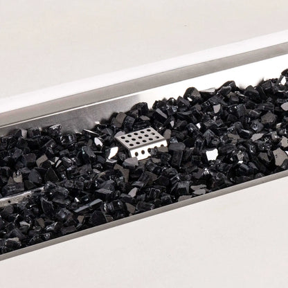 Detailed close-up of black reflective fire glass inside the stainless steel burner pan of a white Flame Havens Refuge Series 56 x 30 x 24-inch rectangular propane gas GFRC concrete fire pit, with the perforated ignition cage visible near the center.