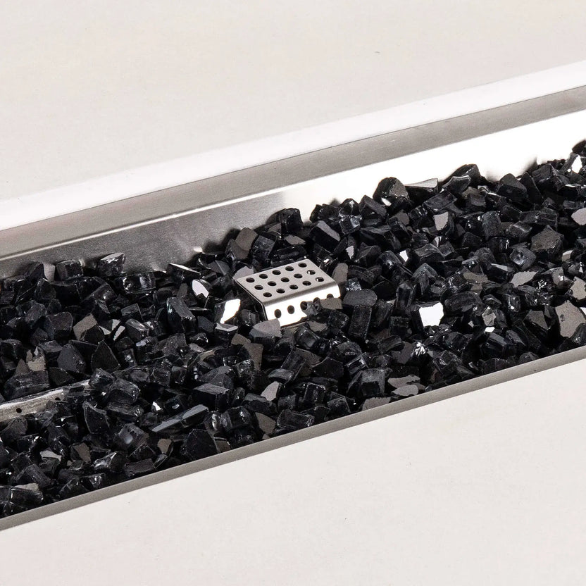 Detailed close-up of black reflective fire glass inside the stainless steel burner pan of a white Flame Havens Refuge Series 56 x 30 x 24-inch rectangular propane gas GFRC concrete fire pit, with the perforated ignition cage visible near the center.