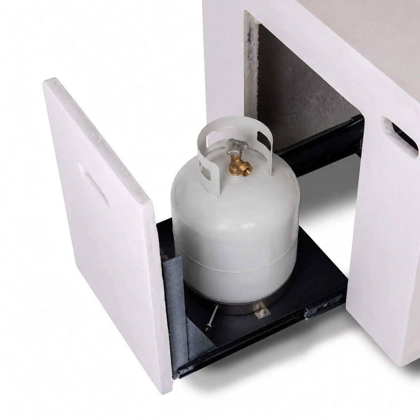 Open hidden propane tank drawer on the white Flame Havens Refuge Series 56 x 30 x 24-inch rectangular GFRC concrete fire pit, shown with a standard propane cylinder positioned securely on the black slide-out platform for convenient access.