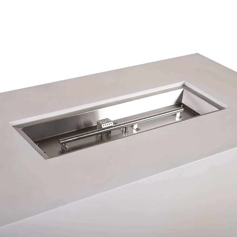Angled close-up of the stainless steel burner assembly inside a white Flame Havens Refuge Series 56 x 30 x 24-inch rectangular propane gas fire pit, showing the linear burner tube, mounting brackets, polished burner pan, gas fittings, and perforated CSA ignition cage.