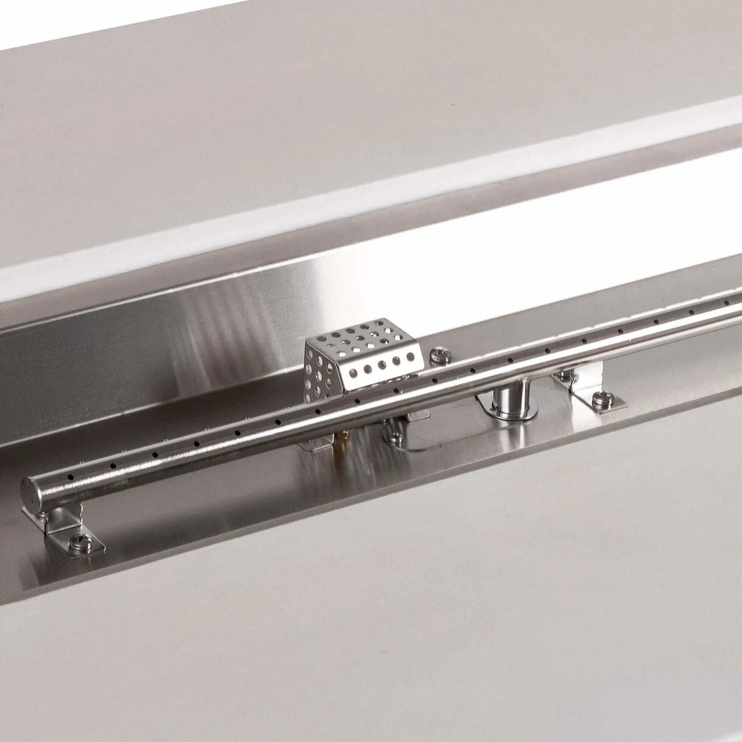 Detailed view of the stainless steel linear burner and CSA ignition components in a white Flame Havens Refuge Series 56 x 30 x 24-inch rectangular propane gas fire pit, highlighting the burner ports, polished pan, mounting hardware, gas inlet, and perforated ignition cage.