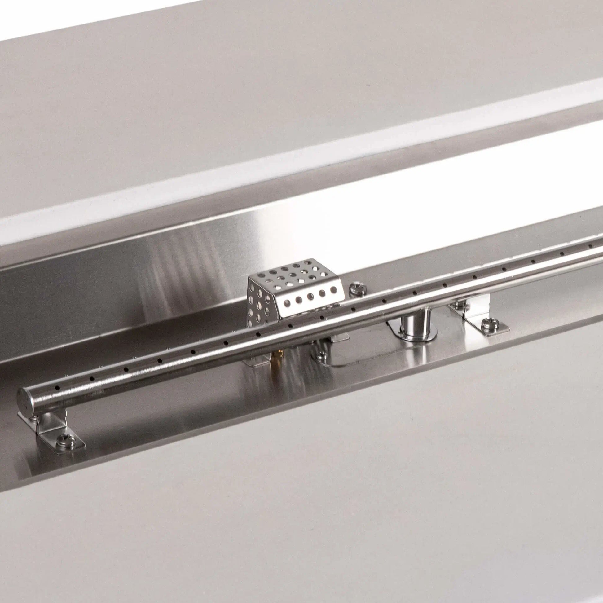 Detailed view of the stainless steel linear burner and CSA ignition components in a white Flame Havens Refuge Series 56 x 30 x 24-inch rectangular propane gas fire pit, highlighting the burner ports, polished pan, mounting hardware, gas inlet, and perforated ignition cage.