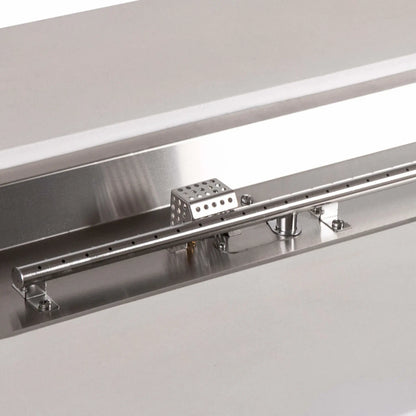 Detailed view of the stainless steel linear burner and CSA ignition components in a white Flame Havens Refuge Series 56 x 30 x 24-inch rectangular propane gas fire pit, highlighting the burner ports, polished pan, mounting hardware, gas inlet, and perforated ignition cage.