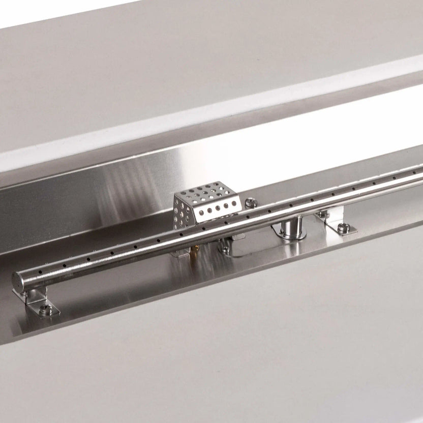 Detailed view of the stainless steel linear burner and CSA ignition components in a white Flame Havens Refuge Series 56 x 30 x 24-inch rectangular propane gas fire pit, highlighting the burner ports, polished pan, mounting hardware, gas inlet, and perforated ignition cage.