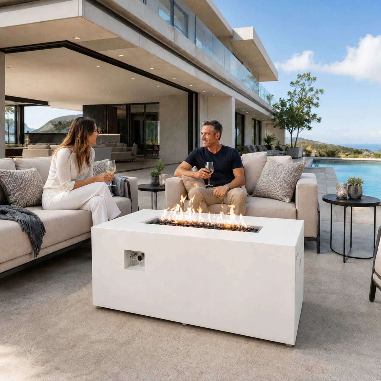 White Flame Havens Refuge Series 56 x 30 x 24-inch rectangular propane gas fire pit with tempered black fire glass and visible controls, burning between two adults enjoying wine beside a pool on the patio of a modern luxury home.
