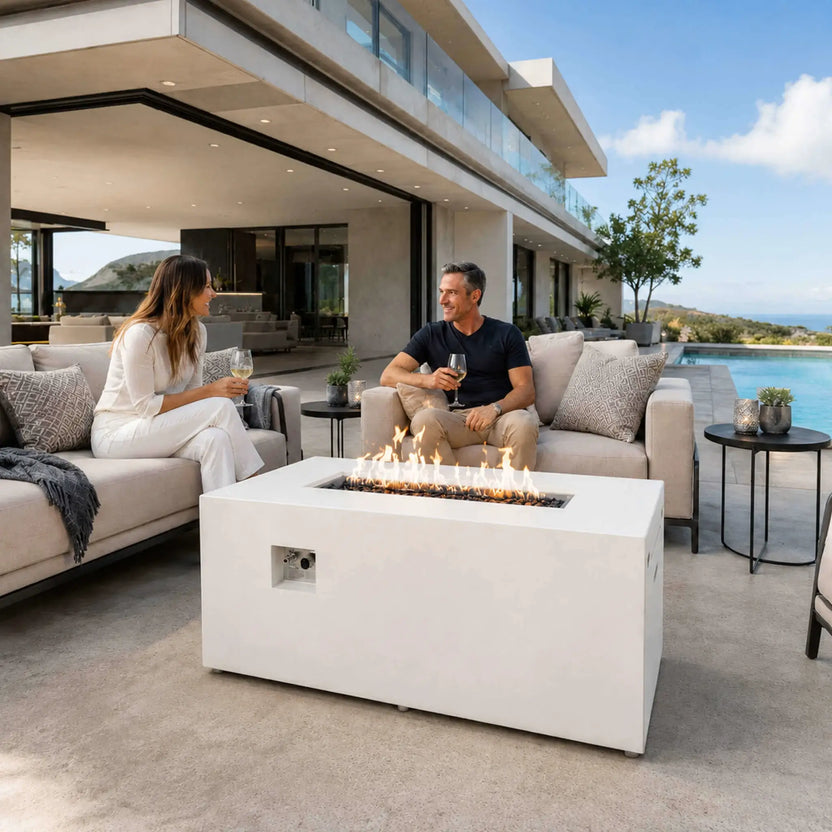 White Flame Havens Refuge Series 56 x 30 x 24-inch rectangular propane gas fire pit with tempered black fire glass and visible controls, burning between two adults enjoying wine beside a pool on the patio of a modern luxury home.