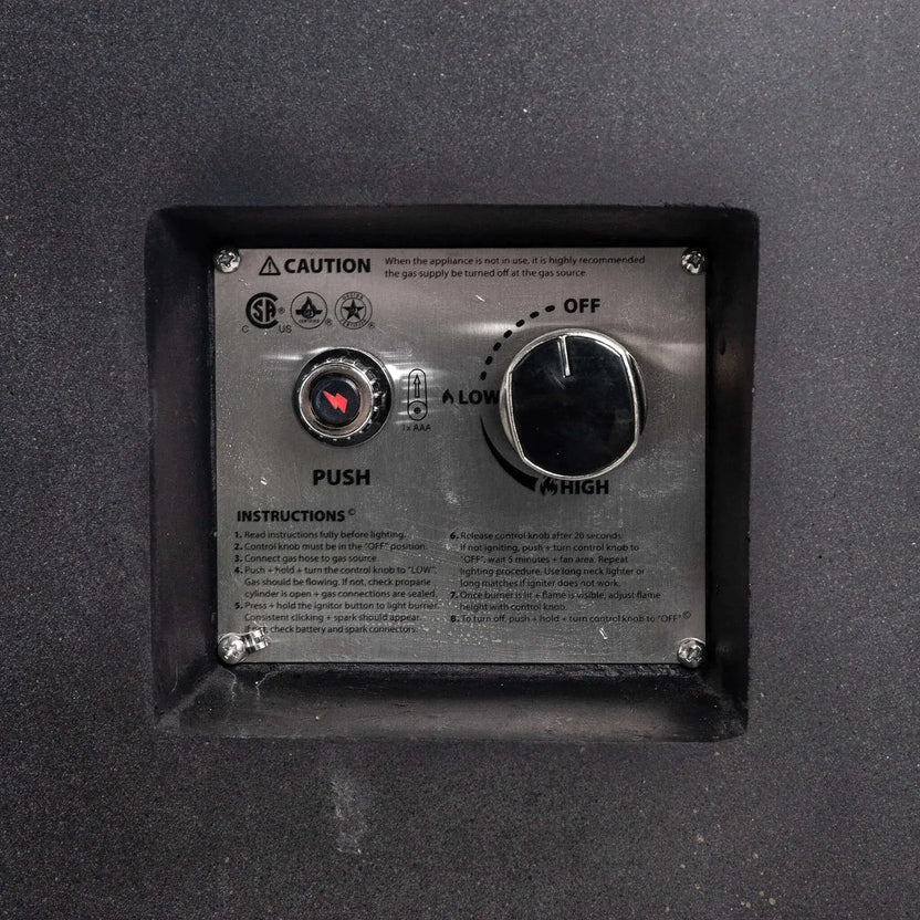 Close-up of the recessed CSA ignition controls on the black Flame Havens Refuge Series rectangular GFRC concrete propane fire pit. The stainless steel panel includes a push-button spark igniter, AAA battery indicator, adjustable control knob with off, low, and high settings, safety notice, and lighting instructions.
