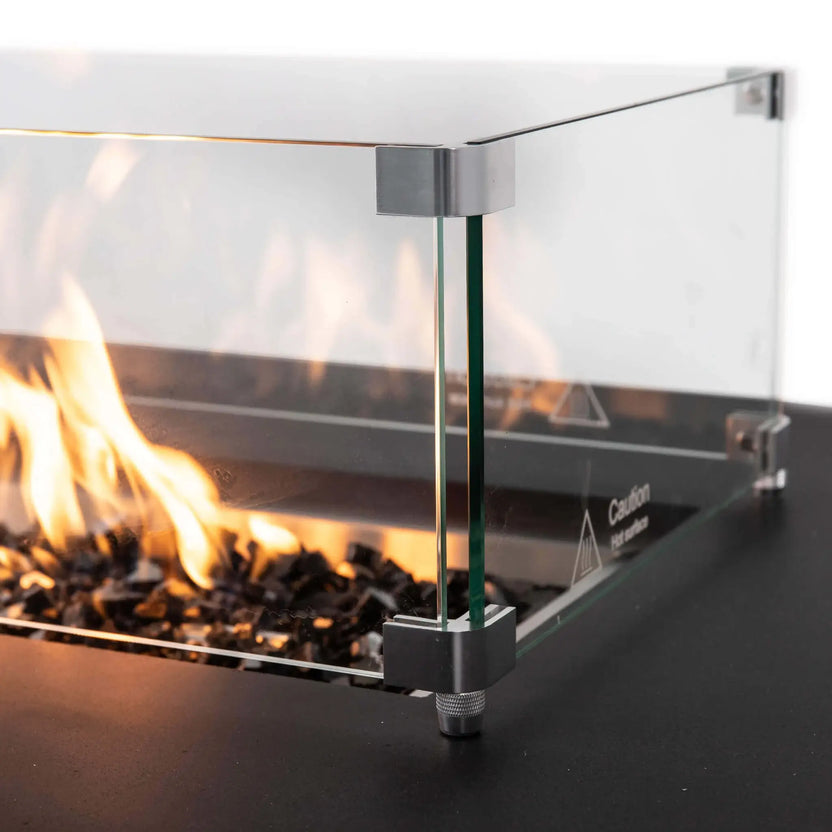 Close-up of the tempered glass wind guard surrounding the flames on the black Flame Havens Refuge Series 56 x 30 x 24-inch rectangular propane fire pit. The image highlights the clear glass panels, stainless steel corner brackets, adjustable foot, tempered black fire glass, and burning flame.
