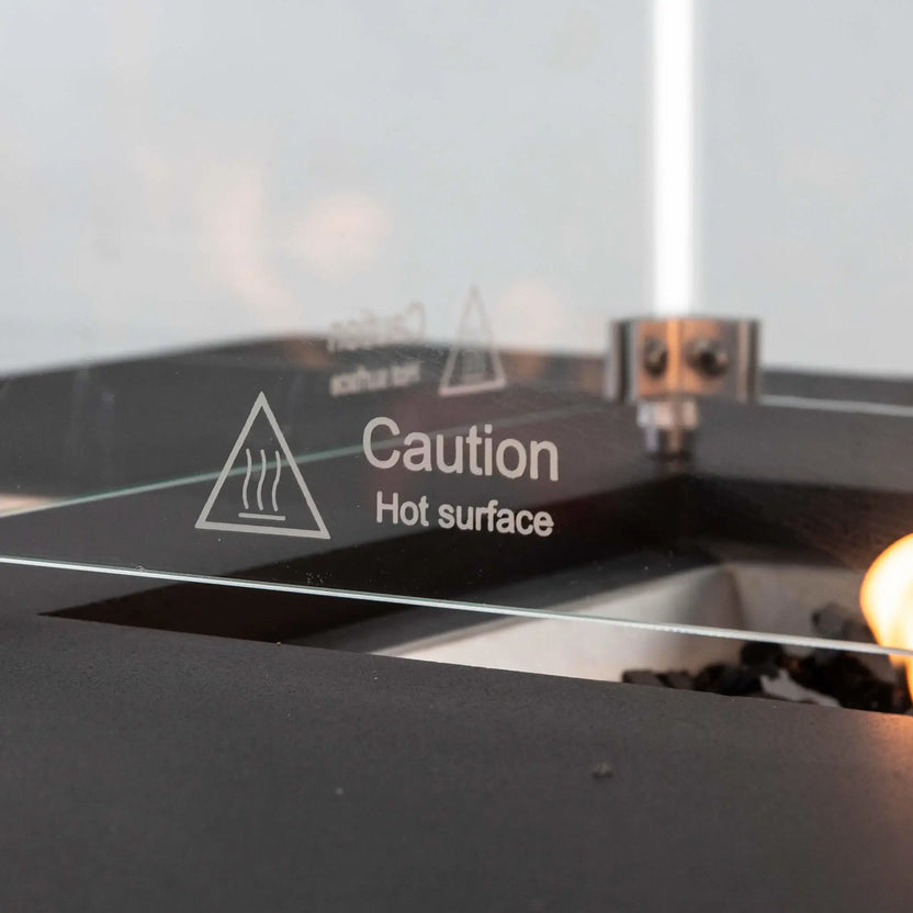 Detailed close-up of the tempered glass wind guard on the black Flame Havens Refuge Series rectangular GFRC propane fire pit. A clearly printed “Caution Hot Surface” warning appears on the glass, with stainless steel corner hardware, black fire glass, and part of the active flame visible.
