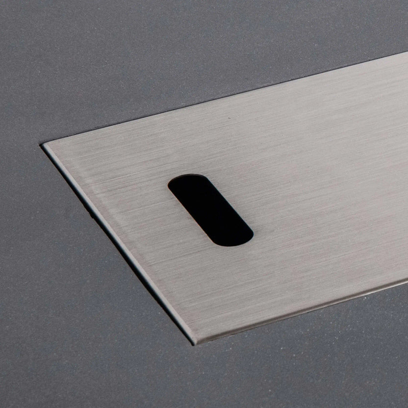Close-up of the brushed stainless steel burner cover fitted into the Flame Havens Refuge Series 56 x 30 x 24-inch rectangular gray propane fire pit. The image highlights the cover’s smooth metal grain, clean flush-mounted edges and rounded rectangular handle opening against the textured GFRC concrete surface.