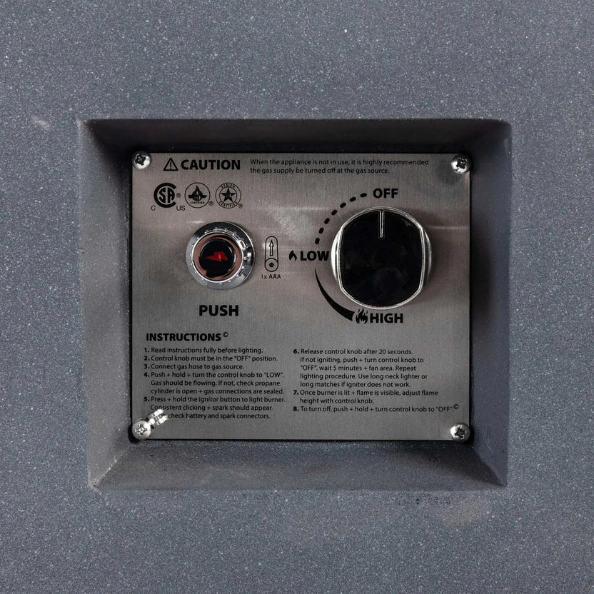 Close-up of the recessed CSA ignition controls on the Flame Havens Refuge Series 56 x 30 x 24-inch gray rectangular propane fire pit. The stainless steel panel includes a push-button spark igniter, AAA battery indicator, large flame adjustment knob marked Off, Low and High, certification symbols, caution notice and lighting instructions.