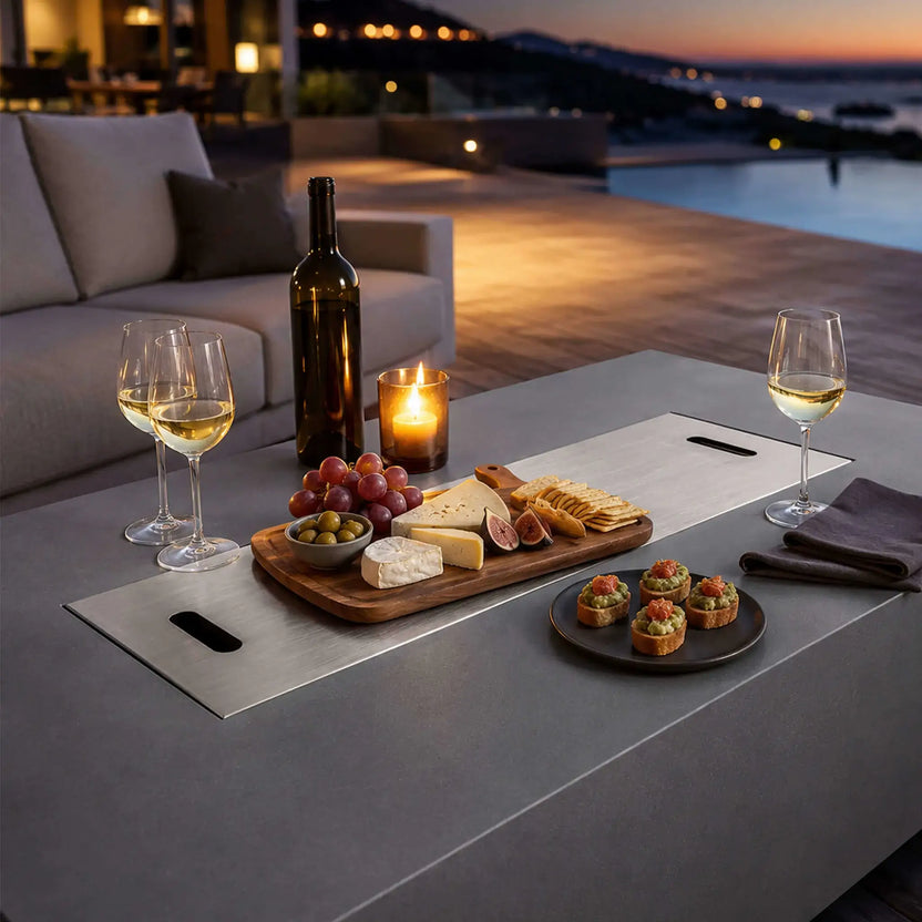 Flame Havens Refuge Series 56 x 30 x 24-inch gray propane fire pit used as a patio table with its stainless steel burner cover installed. Wine glasses, a bottle, candle, cheese board, grapes, olives, crackers and appetizers are arranged across the covered surface beside outdoor seating and a pool at dusk.