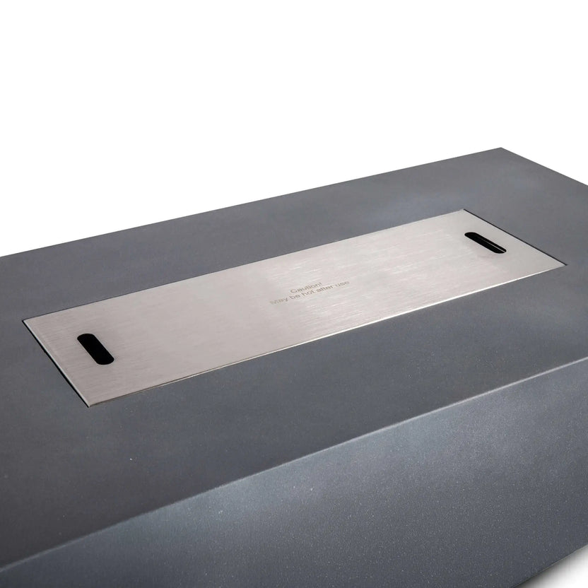 Angled view of the stainless steel burner cover installed flush in the Flame Havens Refuge Series 56 x 30 x 24-inch gray GFRC propane fire pit. The long brushed metal cover conceals the burner opening and features two recessed handles with a centered caution message noting the surface may remain hot after use.