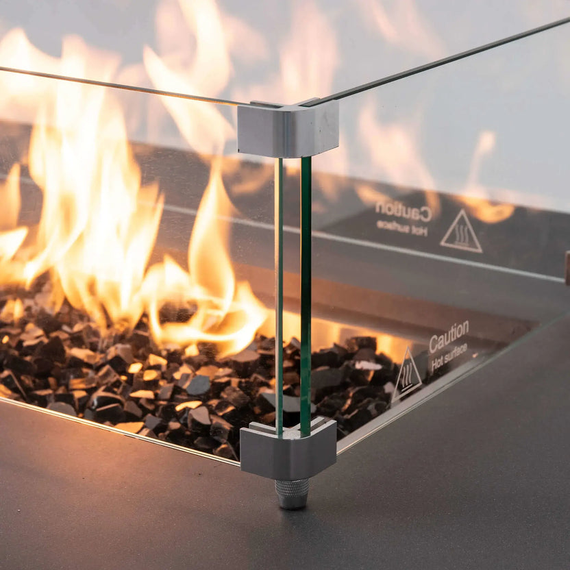 Close-up of the tempered glass wind guard surrounding the lit burner on the Flame Havens Refuge Series 56 x 30 x 24-inch gray propane fire pit. Bright flames rise from tempered black fire glass behind clear panels secured by metal corner brackets, with the lower mounting foot and hot-surface caution markings visible.