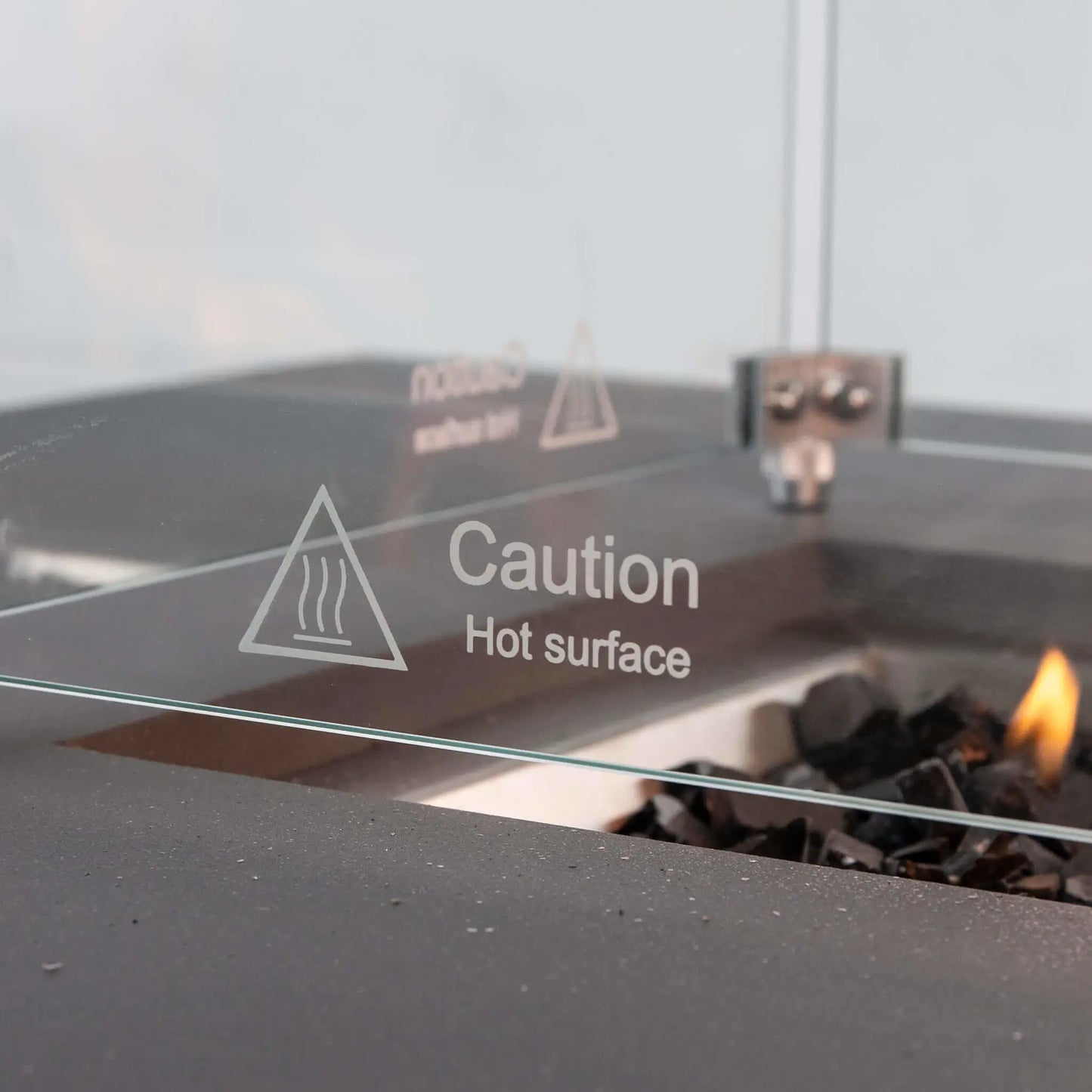Detailed view of the “Caution Hot Surface” marking etched into the tempered glass wind guard on the Flame Havens Refuge Series 56 x 30 x 24-inch gray propane fire pit. The clear panel, metal corner hardware, textured GFRC ledge, tempered black fire glass and a small active flame are visible in the background.