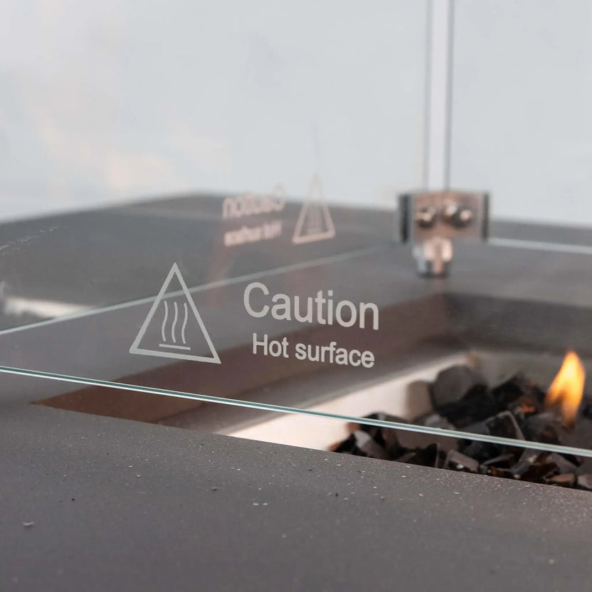 Detailed view of the “Caution Hot Surface” marking etched into the tempered glass wind guard on the Flame Havens Refuge Series 56 x 30 x 24-inch gray propane fire pit. The clear panel, metal corner hardware, textured GFRC ledge, tempered black fire glass and a small active flame are visible in the background.