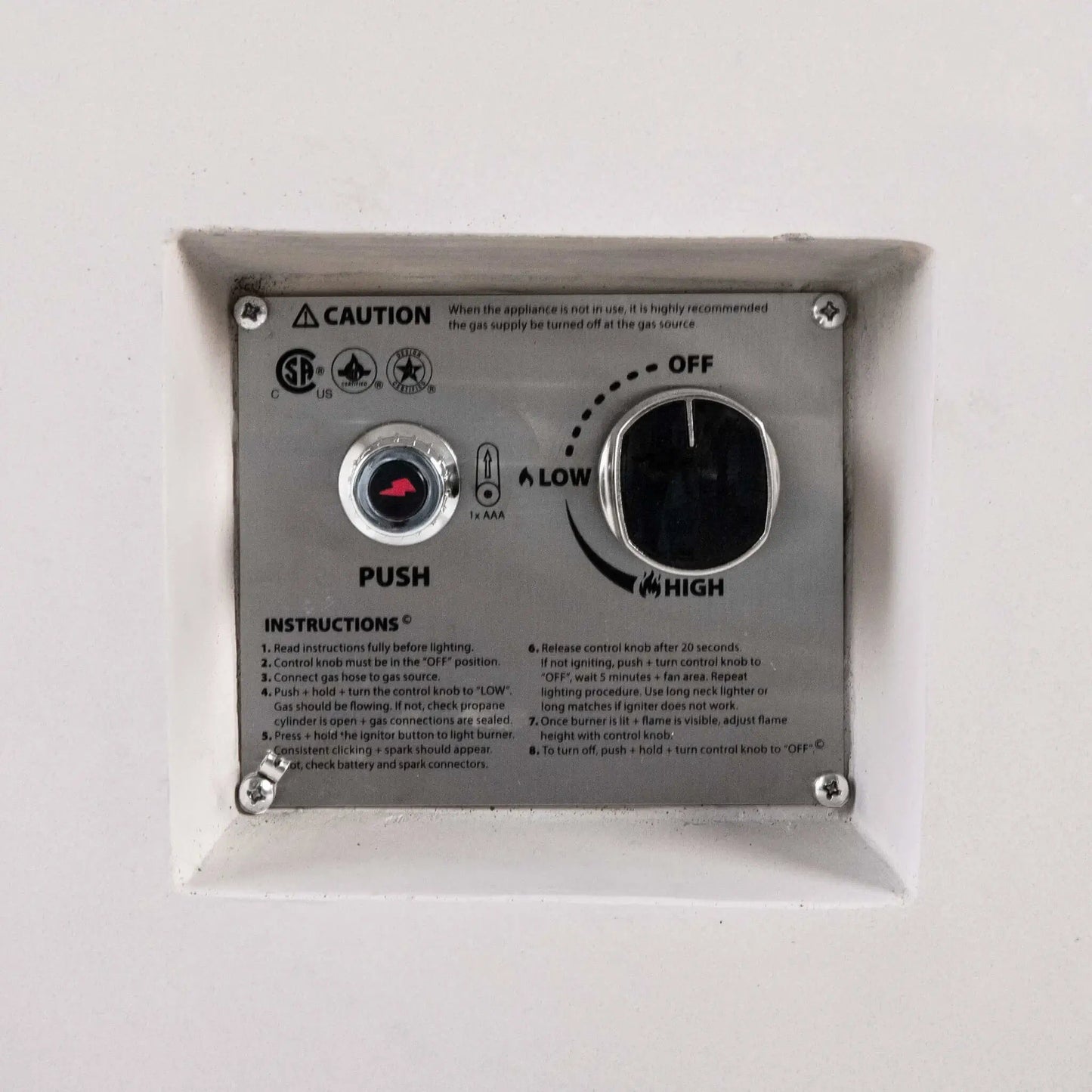Close-up of the recessed CSA ignition controls on a white Flame Havens Refuge Series 56 x 30 x 24-inch rectangular propane gas fire pit, showing the push-button spark igniter, AAA battery indicator, OFF-to-LOW-to-HIGH flame control knob, safety certifications, caution notice, and lighting instructions.