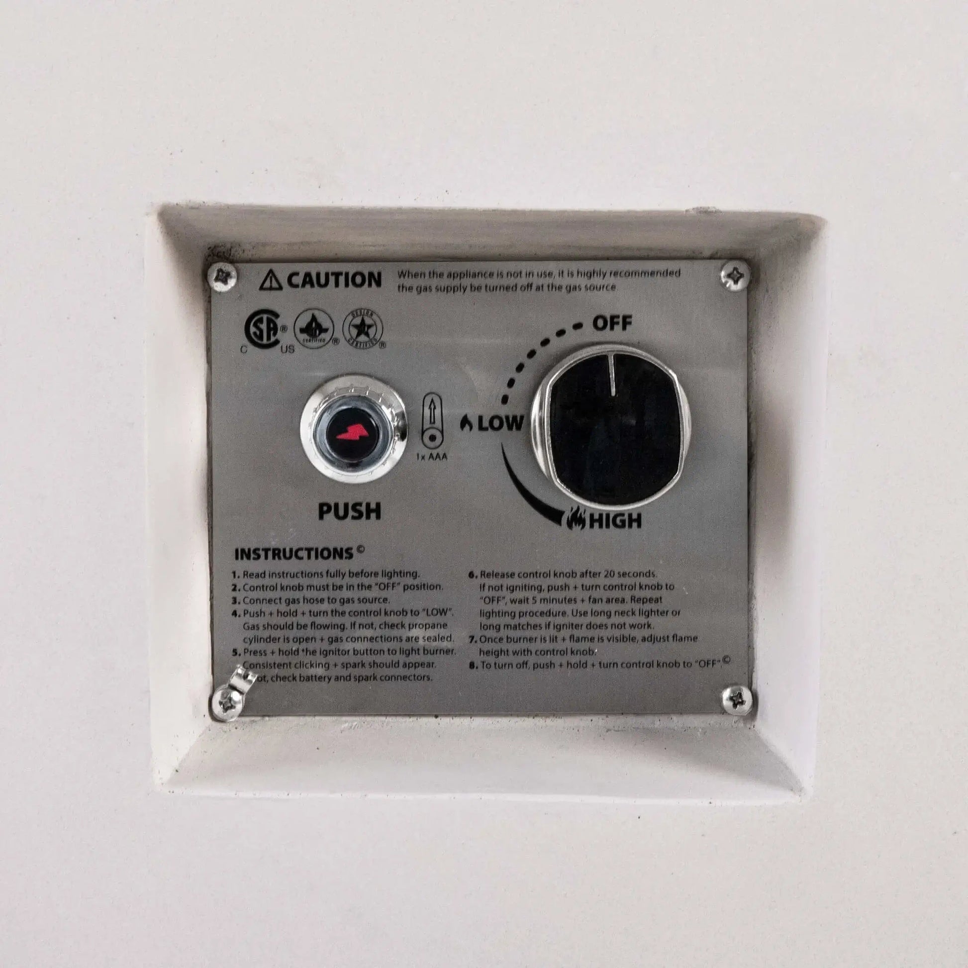 Close-up of the recessed CSA ignition controls on a white Flame Havens Refuge Series 56 x 30 x 24-inch rectangular propane gas fire pit, showing the push-button spark igniter, AAA battery indicator, OFF-to-LOW-to-HIGH flame control knob, safety certifications, caution notice, and lighting instructions.