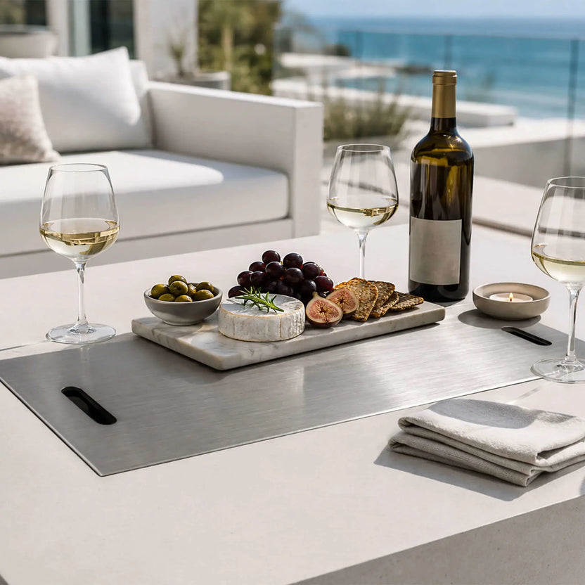 Flame Havens Refuge Series 56 x 30 x 24-inch white rectangular propane gas fire pit fitted with a stainless steel burner cover and used as an outdoor serving table, arranged with wine, glasses, cheese, grapes, figs, olives, crackers, and a candle on a coastal patio.
