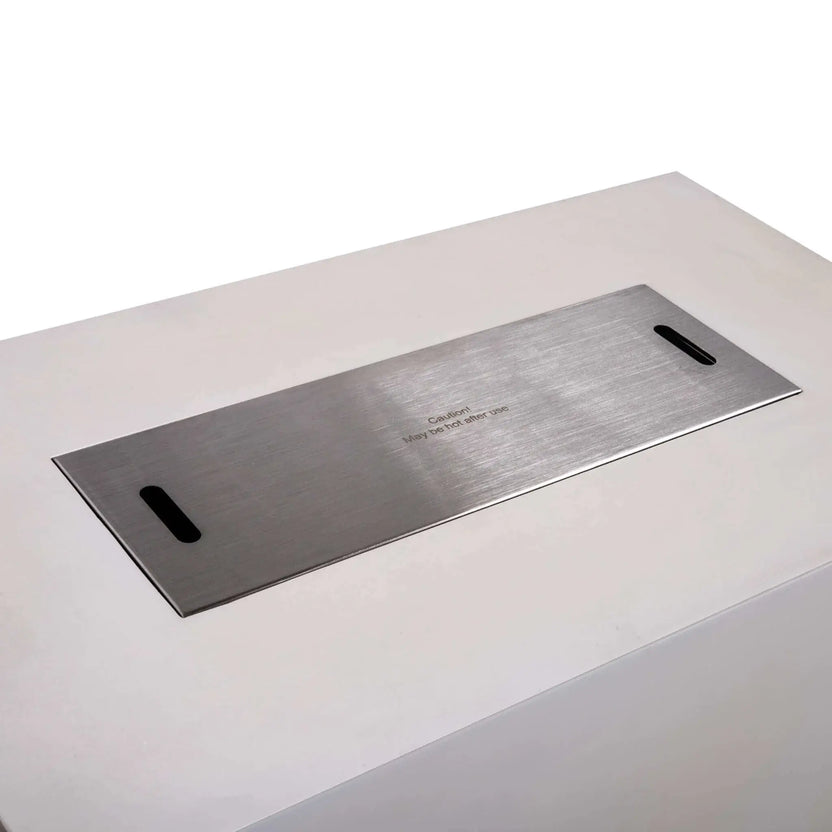 Brushed stainless steel burner cover installed over the opening of a white Flame Havens Refuge Series 56 x 30 x 24-inch rectangular propane gas fire pit, featuring two recessed lifting slots and a centered caution message indicating the surface may be hot after use.