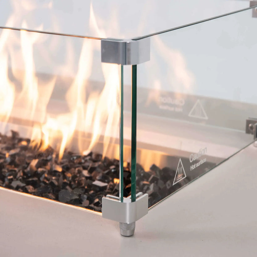 Close-up of the tempered glass wind guard on a white Flame Havens Refuge Series 56 x 30 x 24-inch rectangular propane gas fire pit, showing bright flames over tempered black fire glass, clear glass panels, polished metal corner brackets, raised feet, and a hot-surface caution marking.