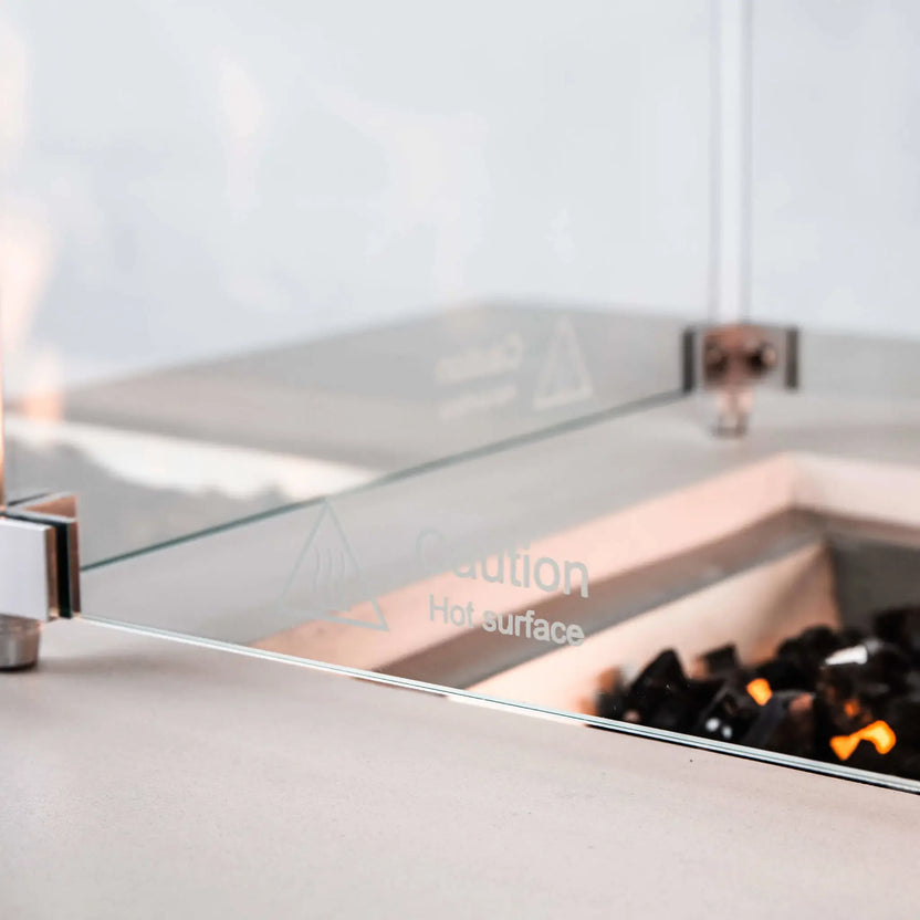 Detailed side view of the tempered glass wind guard on a white Flame Havens Refuge Series 56 x 30 x 24-inch rectangular propane gas fire pit, highlighting the clear glass panel, metal corner hardware, etched “Caution Hot Surface” warning, and glowing flames over tempered black fire glass.