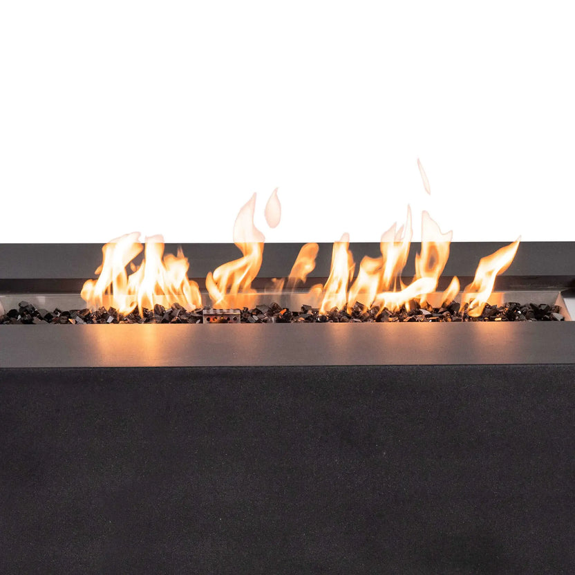 Close-up of bright flames burning across tempered black fire glass in the Flame Havens Refuge Series 56 x 30 x 24-inch rectangular GFRC propane fire pit. The stainless steel burner pan, protective ignition cage, reflective black glass, and textured black concrete surround are clearly visible.