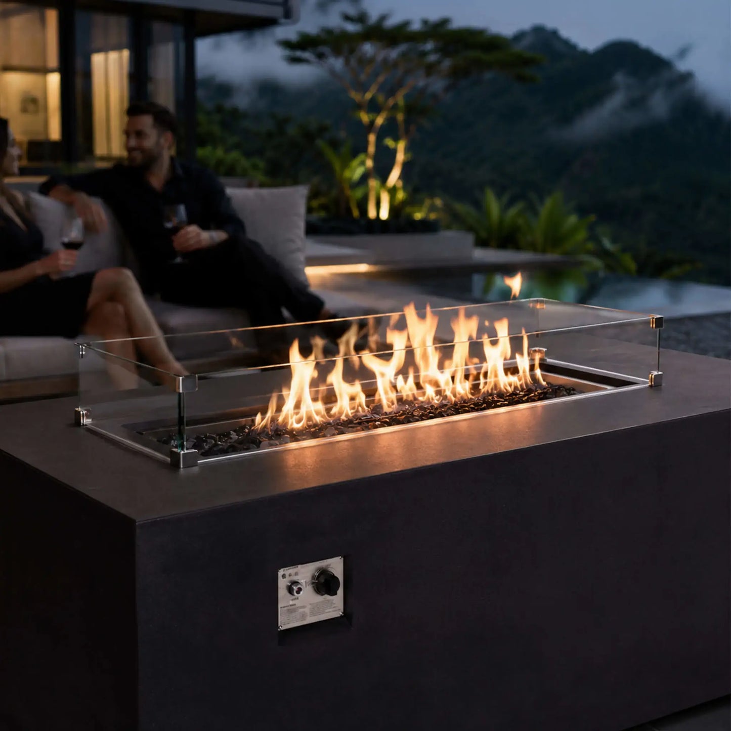 Lifestyle view of the black Flame Havens Refuge Series 56 x 30 x 24-inch rectangular GFRC propane fire pit burning over tempered black fire glass behind a clear wind guard. A couple enjoys wine on a modern patio overlooking a pool, tropical landscaping, and mist-covered mountains at dusk.
