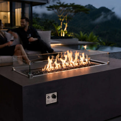 Lifestyle view of the black Flame Havens Refuge Series 56 x 30 x 24-inch rectangular GFRC propane fire pit burning over tempered black fire glass behind a clear wind guard. A couple enjoys wine on a modern patio overlooking a pool, tropical landscaping, and mist-covered mountains at dusk.