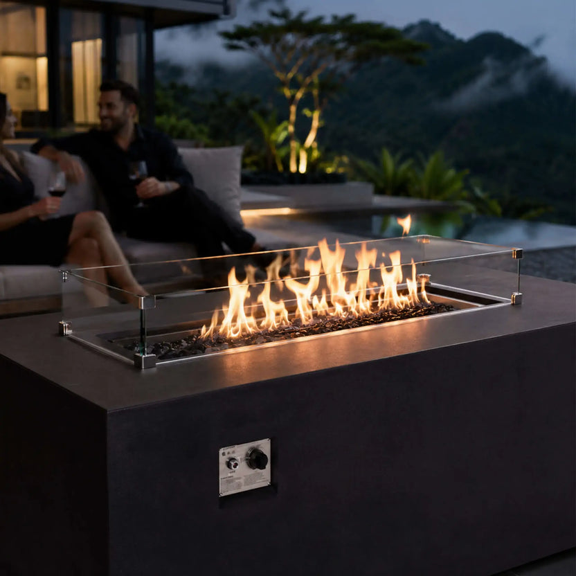 Lifestyle view of the black Flame Havens Refuge Series 56 x 30 x 24-inch rectangular GFRC propane fire pit burning over tempered black fire glass behind a clear wind guard. A couple enjoys wine on a modern patio overlooking a pool, tropical landscaping, and mist-covered mountains at dusk.