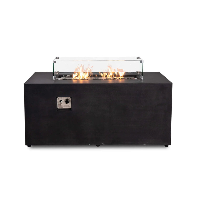 Straight view of the black Flame Havens Refuge Series 56 x 30 x 24-inch rectangular GFRC propane fire pit with an active flame over tempered black fire glass. A clear glass wind guard surrounds the burner and features metal corner brackets, raised feet, and a hot-surface warning.