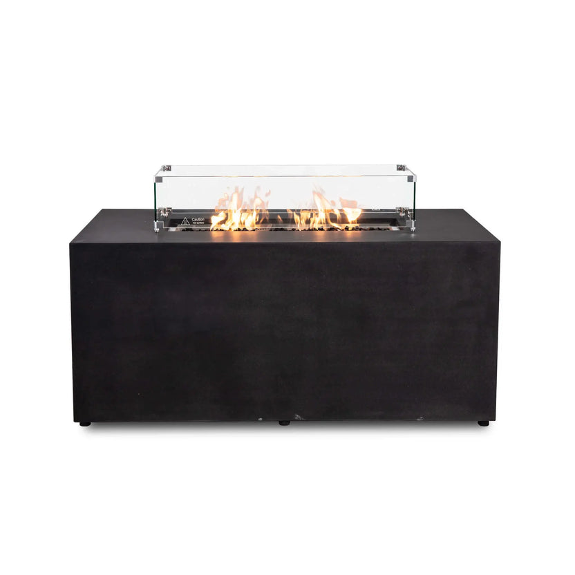 Straight front view of the black Flame Havens Refuge Series 56 x 30 x 24-inch rectangular GFRC concrete propane gas fire pit with flames burning over tempered black fire glass. A clear tempered glass wind guard with stainless steel corner brackets, raised feet, and a hot-surface warning surrounds the active flame.