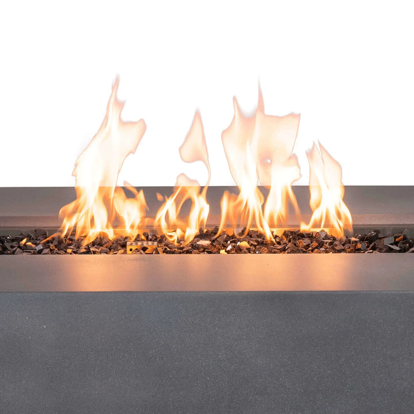 Close-up of bright yellow-orange flames rising from tempered black fire glass in the Flame Havens Refuge Series 56 x 30 x 24-inch gray rectangular propane fire pit. The detailed view shows the reflective glass media, perforated ignition housing, stainless steel burner surround and textured GFRC concrete ledge.