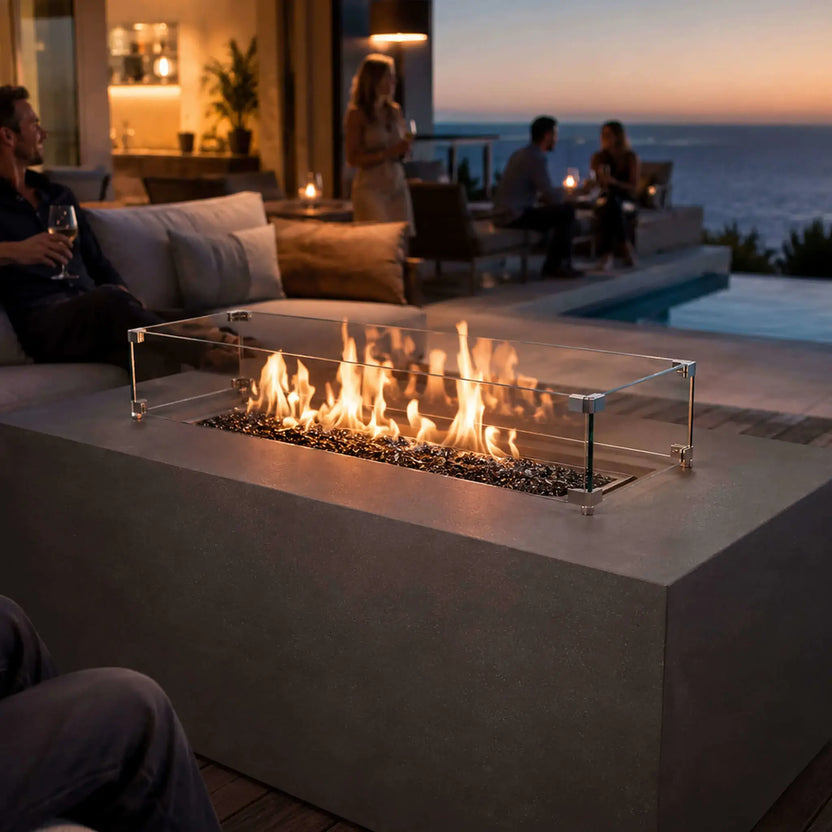 Flame Havens Refuge Series 56 x 30 x 24-inch gray rectangular propane fire pit glowing on a coastal patio at sunset. Flames rise from tempered black fire glass behind a clear wind guard as guests relax with drinks near cushioned seating, a pool and an ocean view, creating an inviting outdoor entertainment setting.