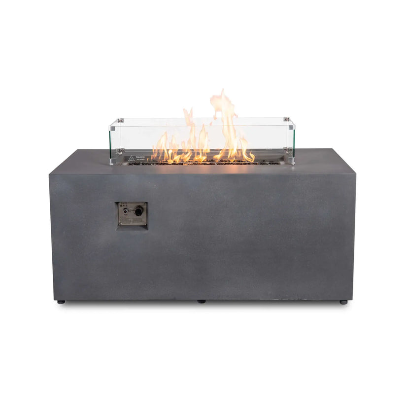 Straight front view of the Flame Havens Refuge Series 56 x 30 x 24-inch gray propane fire pit with a clear tempered glass wind guard surrounding the active flames. Tempered black fire glass fills the linear burner, while metal corner brackets, a hot-surface warning and the recessed front ignition panel are visible.