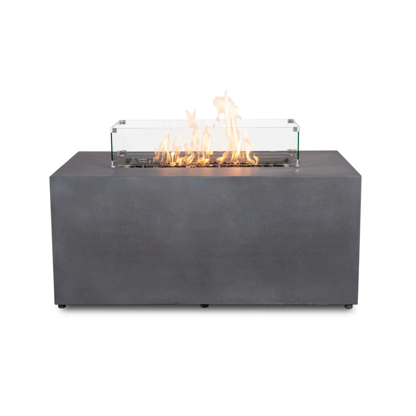 Straight front view of the Flame Havens Refuge Series 56 x 30 x 24-inch rectangular propane gas fire pit in gray GFRC concrete. Bright yellow-orange flames rise from tempered black fire glass behind a clear tempered glass wind guard secured with metal corner brackets. The clean front face and discreet leveling feet are shown against a white background.